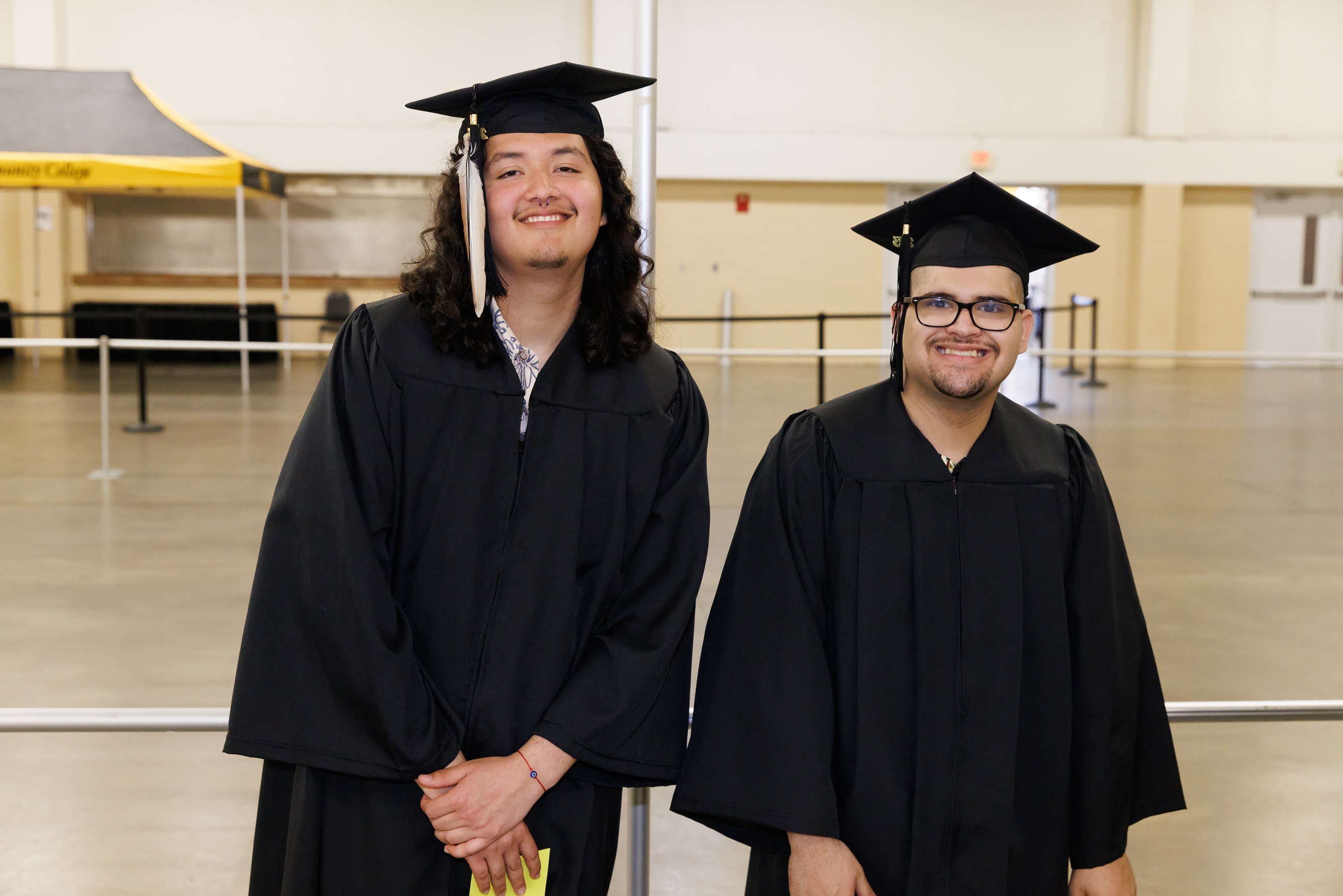 Two graduates pose for a photo. The one on the left has a feather hanging from the tassel on his cap.