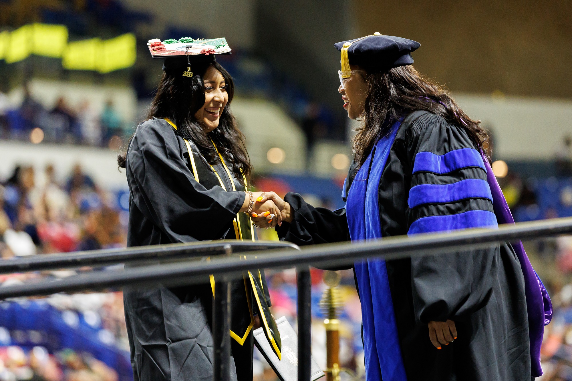A graduate with a decorated cap shakes hands with Dr. Kenjuana McCray, who is dressed in doctoral regalia, on stage.