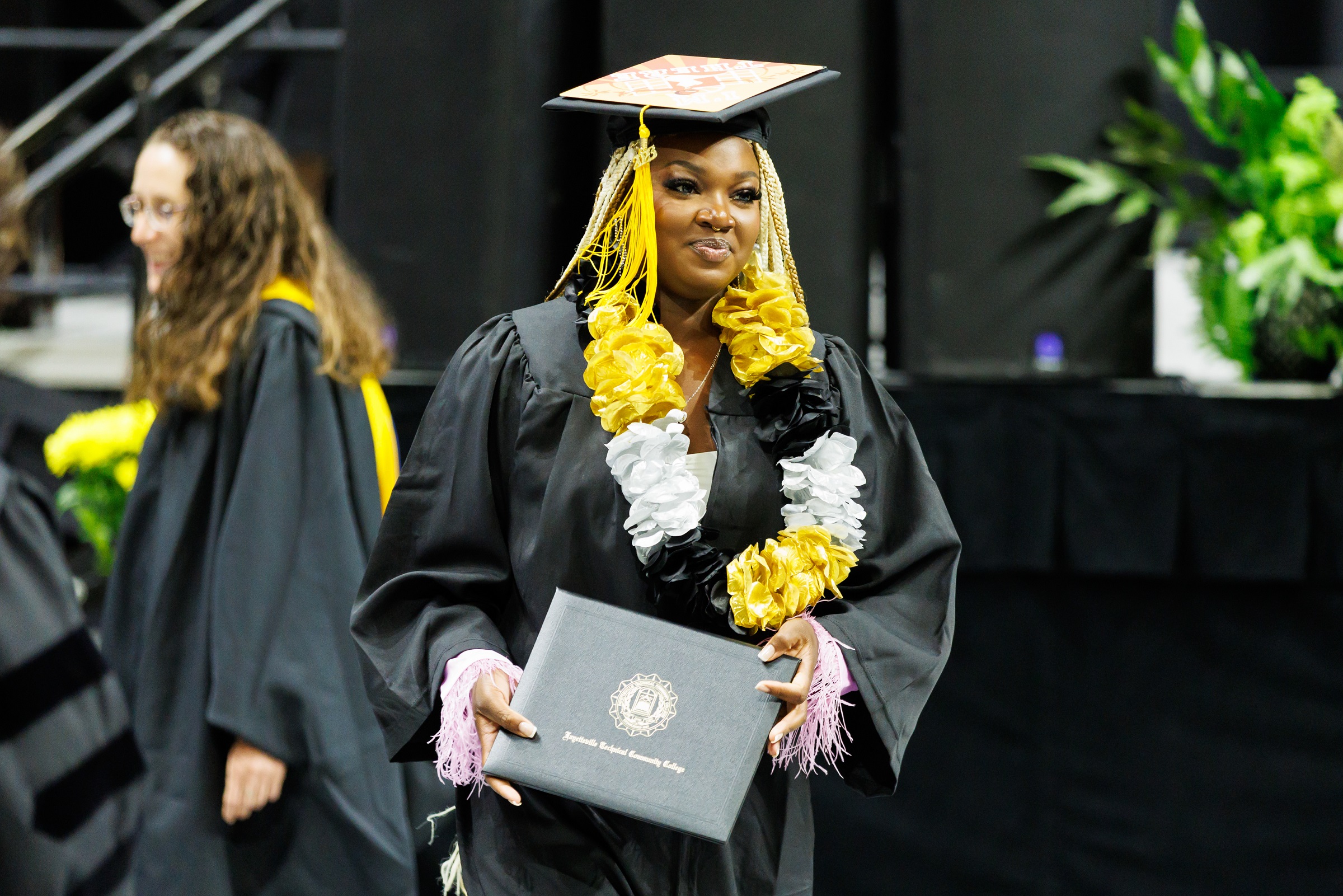 A graduate wearing a gold, white and black lei holds her diploma folder as she walks.