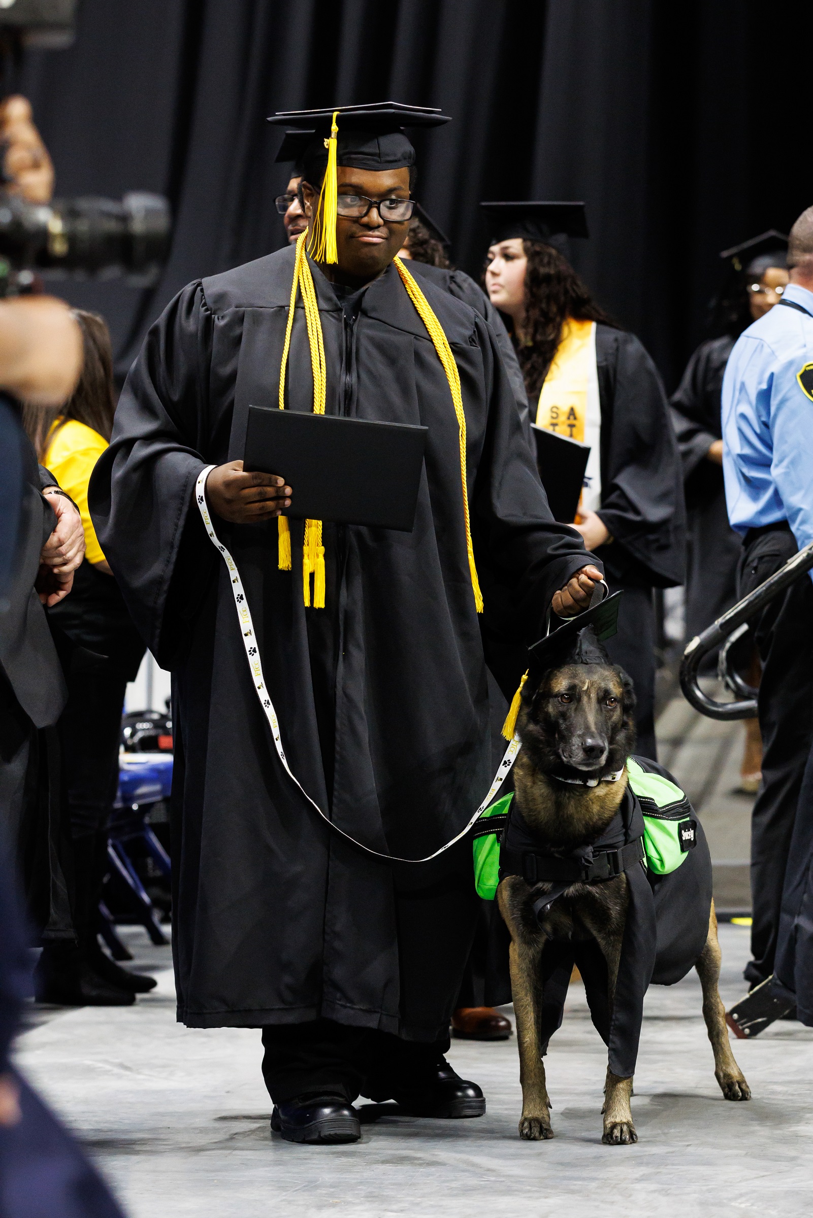 A graduate and his service dog, both dressed in caps and gowns, stand for a photo in front of the stage.