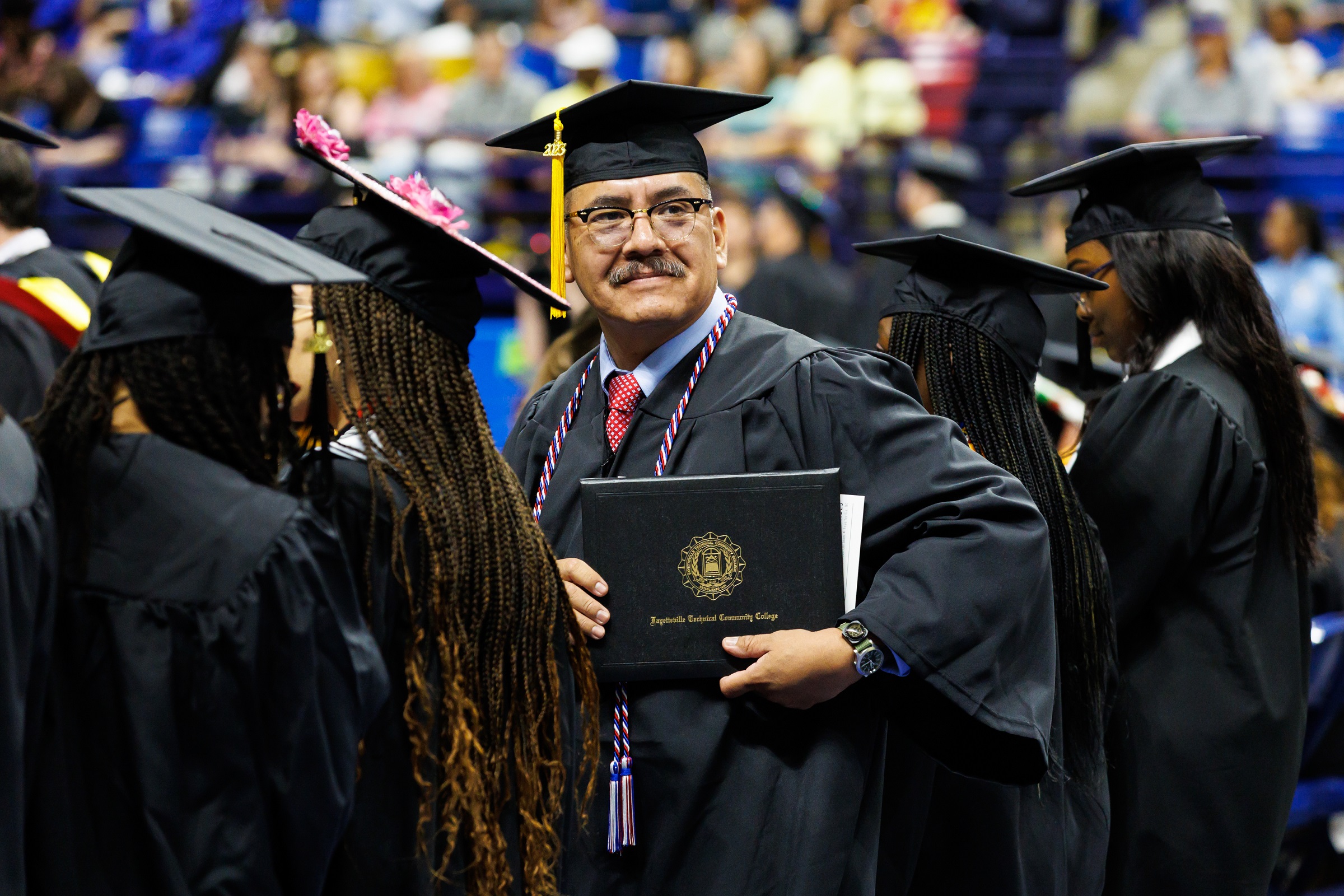 A graduate with a gray moustache and wearing a red, white and blue cord, holds up his diploma folder.