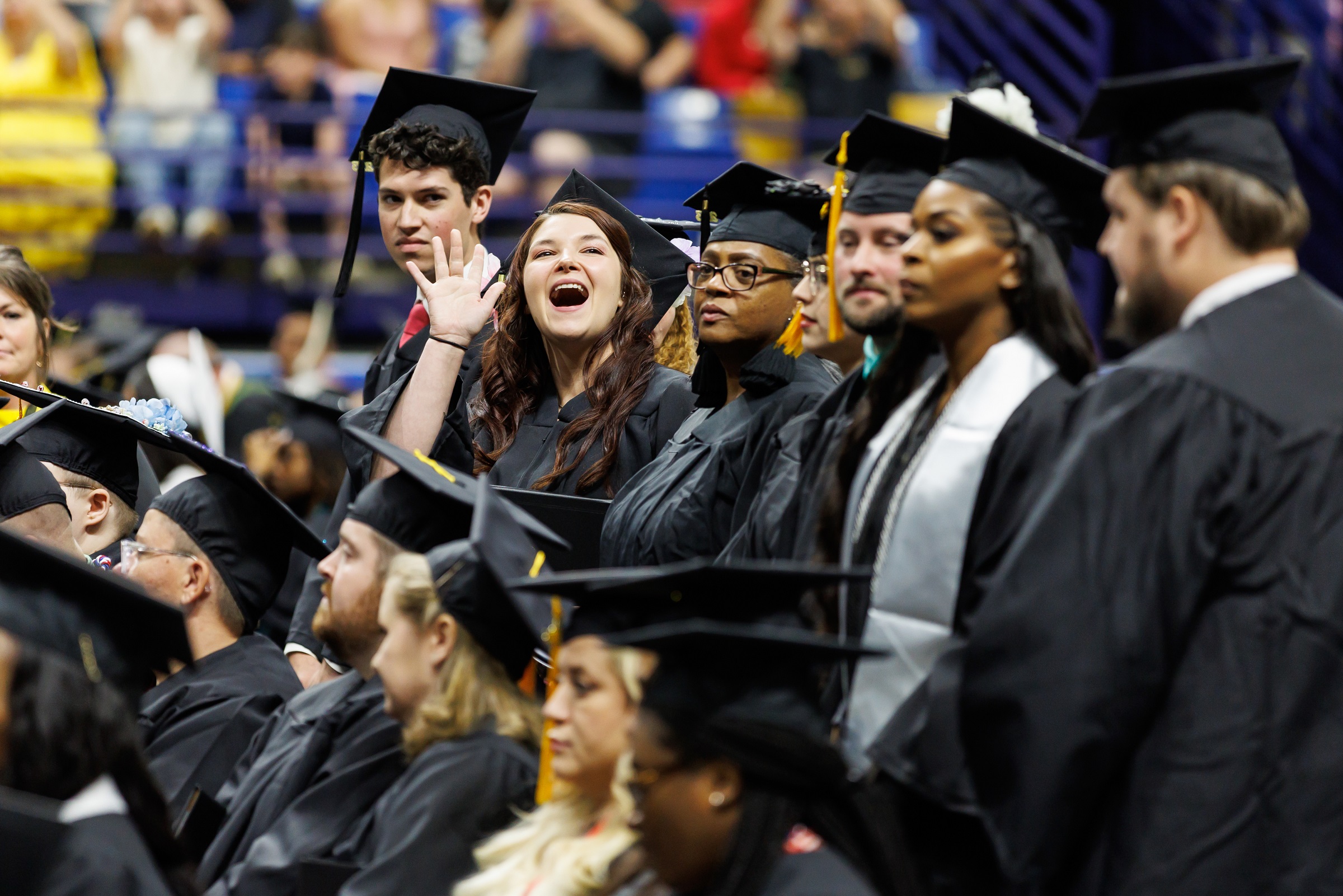A graduate waves to the crowd while she stands in front of her seat.