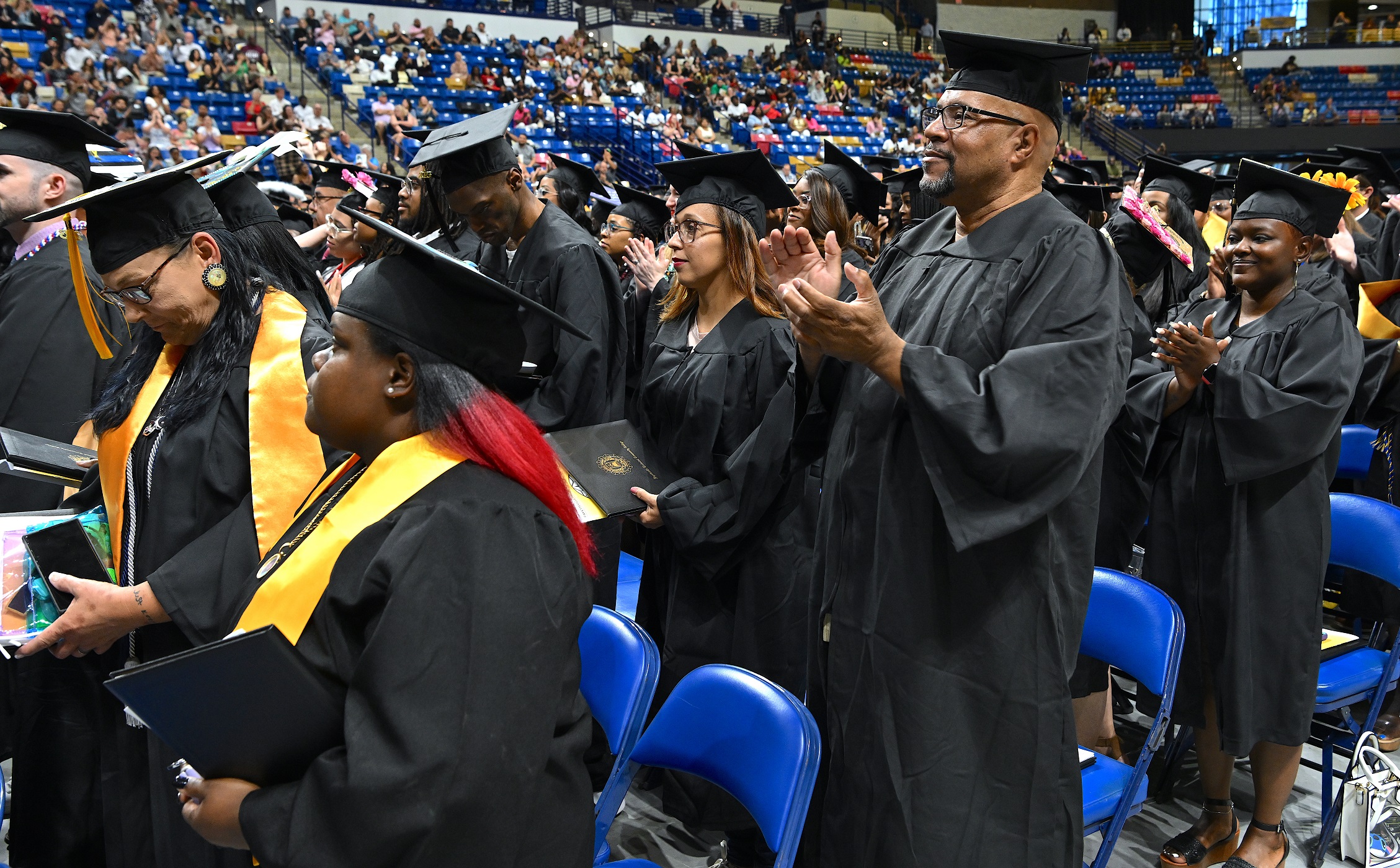 Graduates standing in front of rows of chairs stand and clap.