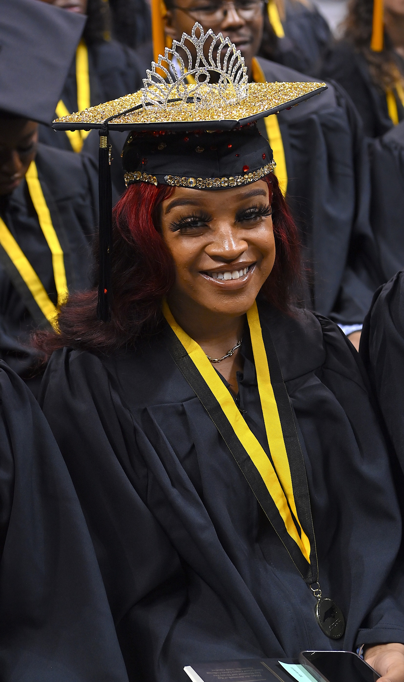 A graduate smiles at the camera from underneath a cap with a tiara attached to the top.